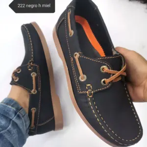 Zapato náutico negro para hombre con cordones camel y suela anaranjada, mostrado en mano y pie con jeans.